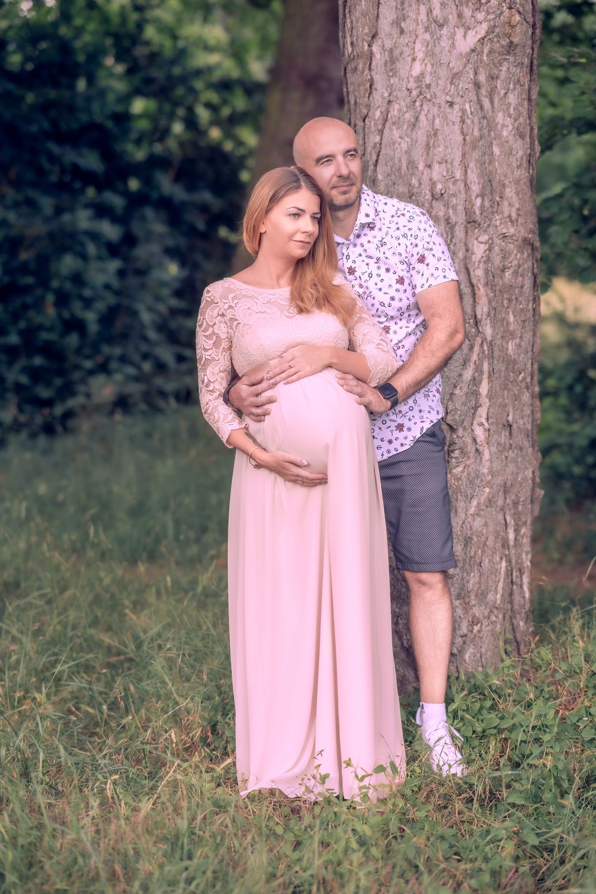 Expectant couple in a pink gown resting against a tree